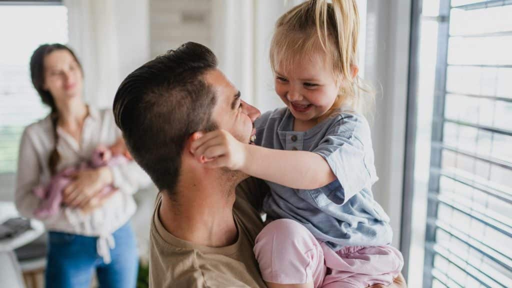 A man holding a laughing little girl while a woman cradles a baby in the background.