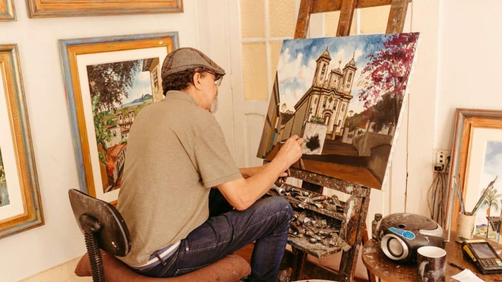 A male artist with a cap paints a building on a canvas in a room.