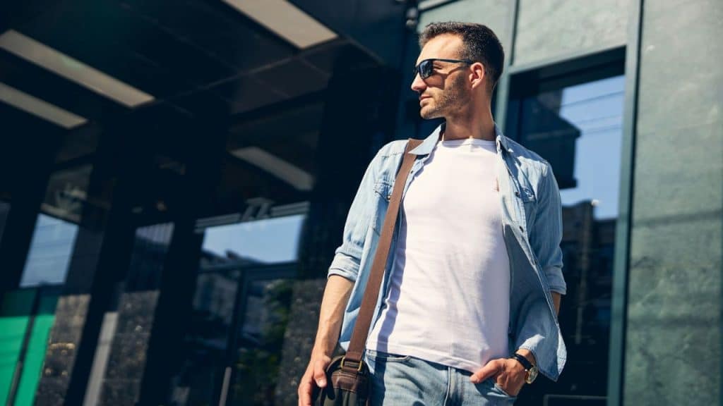 A man in sunglasses, a white t-shirt, and an open denim shirt stands outdoors.