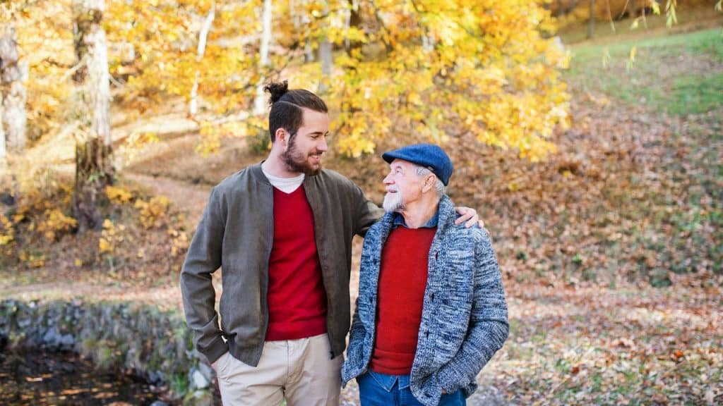 A young man with his arm around an elderly man walking through autumn woods.