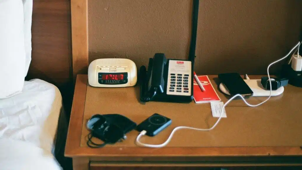 A bedside table with an alarm clock, phone, and charging electronic devices.