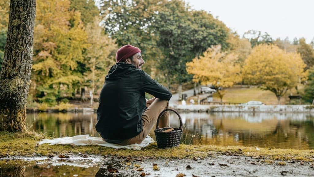 A man in a dark jacket and red beanie sits by a pond with a picnic basket.