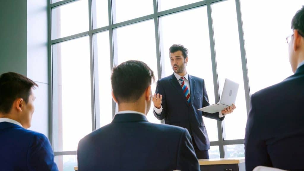 A man in a suit and tie holds a laptop while speaking to an audience.