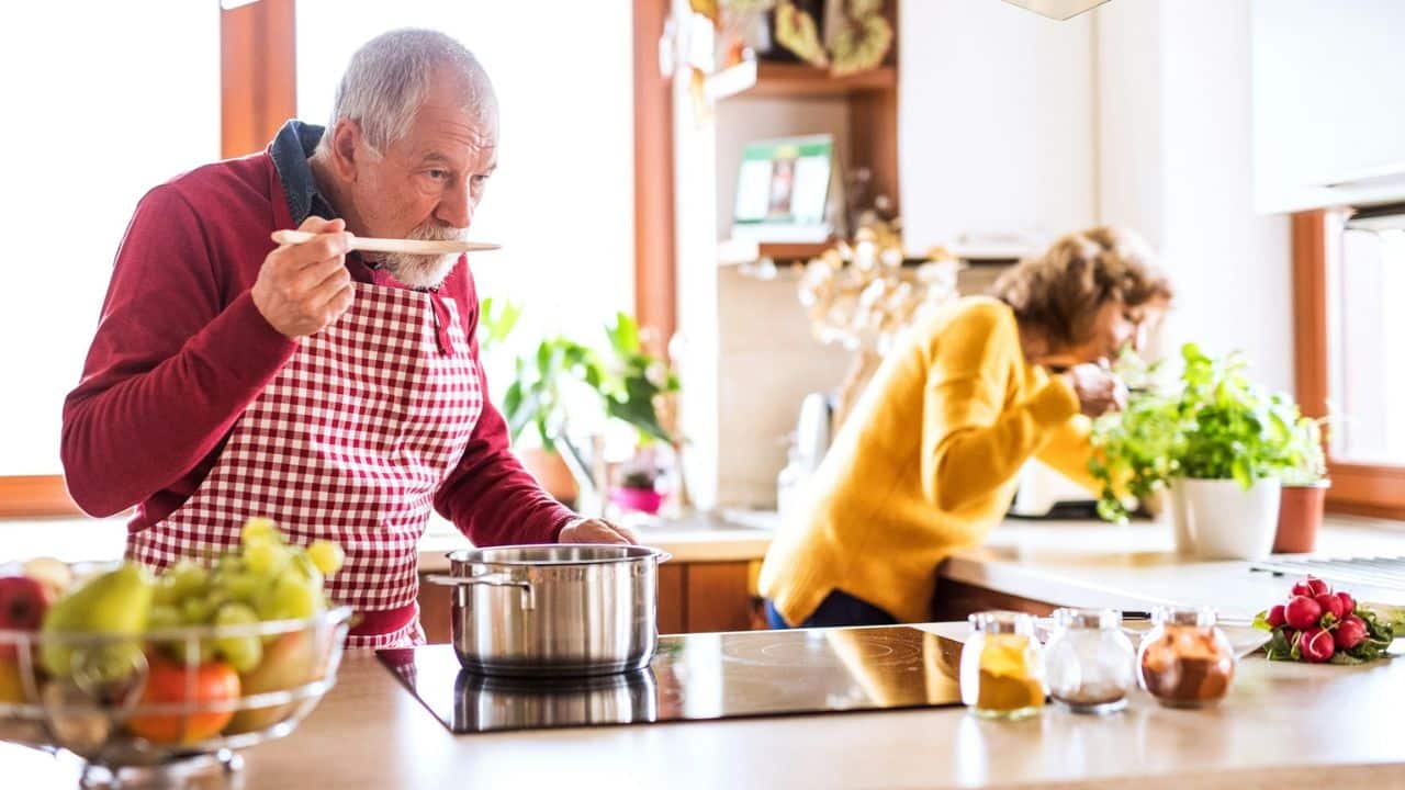 A man in an apron tastes food from a pot while a woman sniffs something in the background.