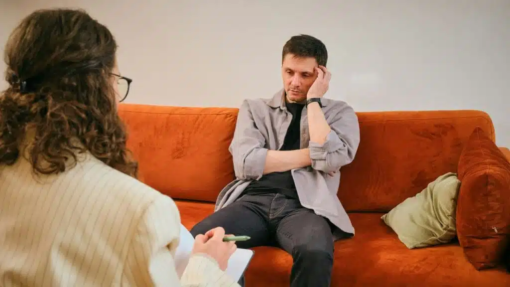 A distressed man sits on a couch while a therapist takes notes during a session.