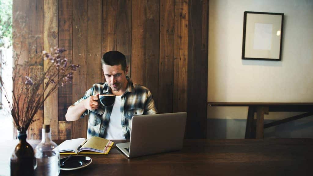 A man drinking coffee at a café table with a laptop and notebook.