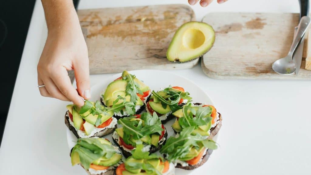 A person adding arugula to rye bread topped with ricotta, tomato, and avocado on a white plate.