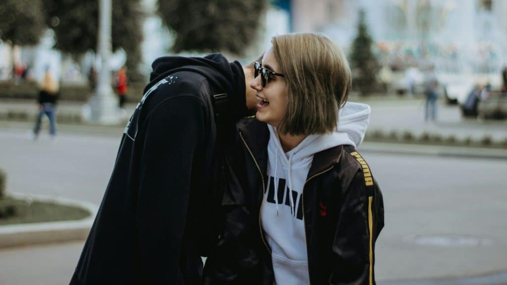 A person whispers into the ear of another person who is laughing outdoors.