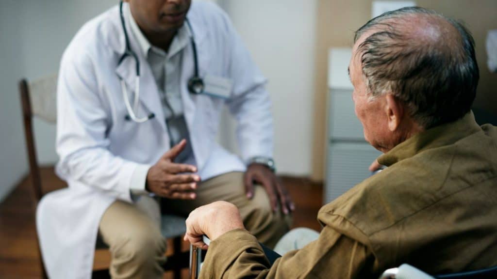 A doctor in a white coat talks to an elderly male patient.