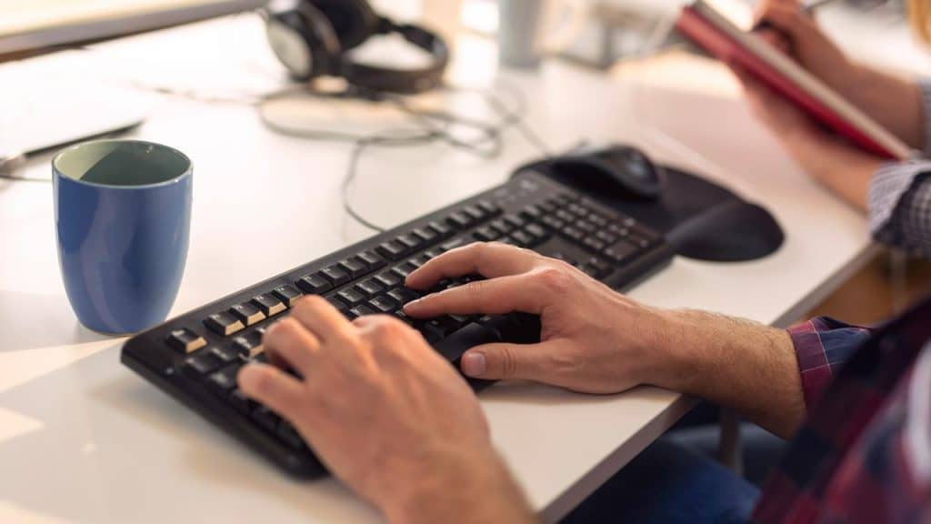 A person typing on a keyboard beside a blue mug.