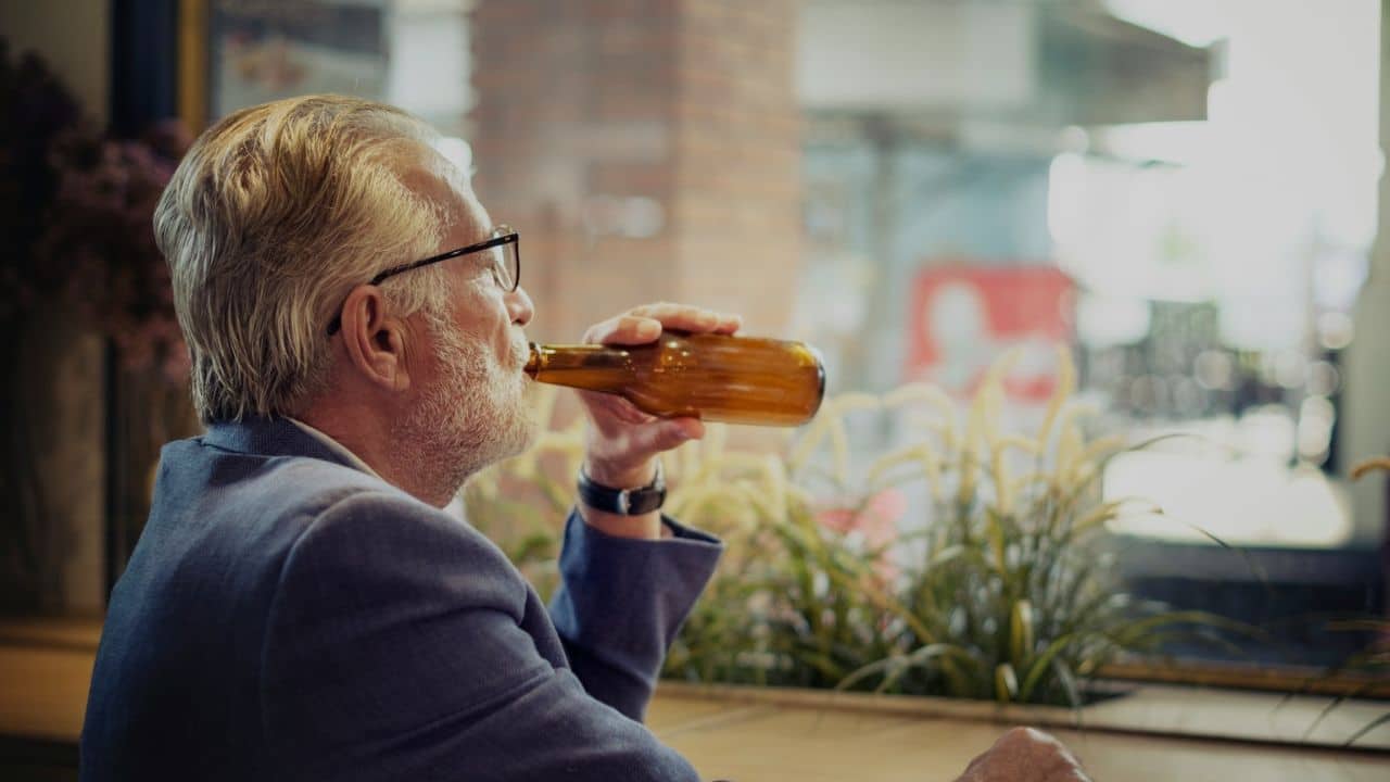 A senior man with a beard and glasses drinks from a brown bottle.