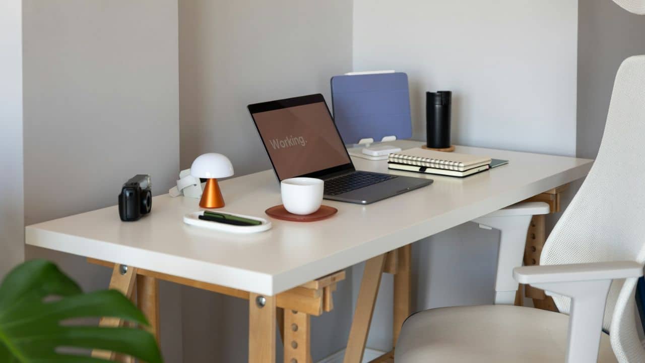 A minimalist workspace features a laptop, tablet, notebook, and coffee mug on a white desk.