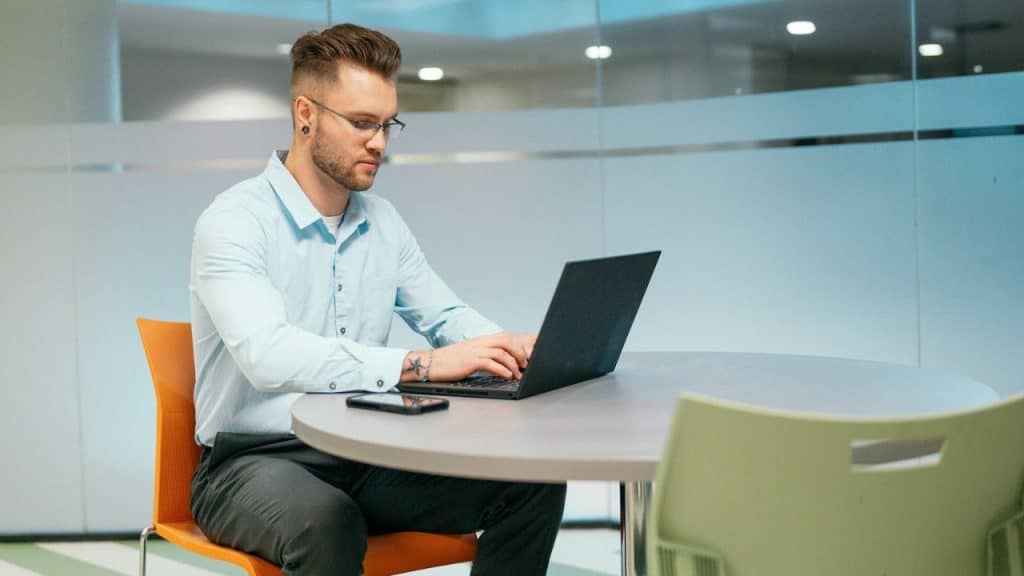A focused man with glasses works on a laptop at a round table.
