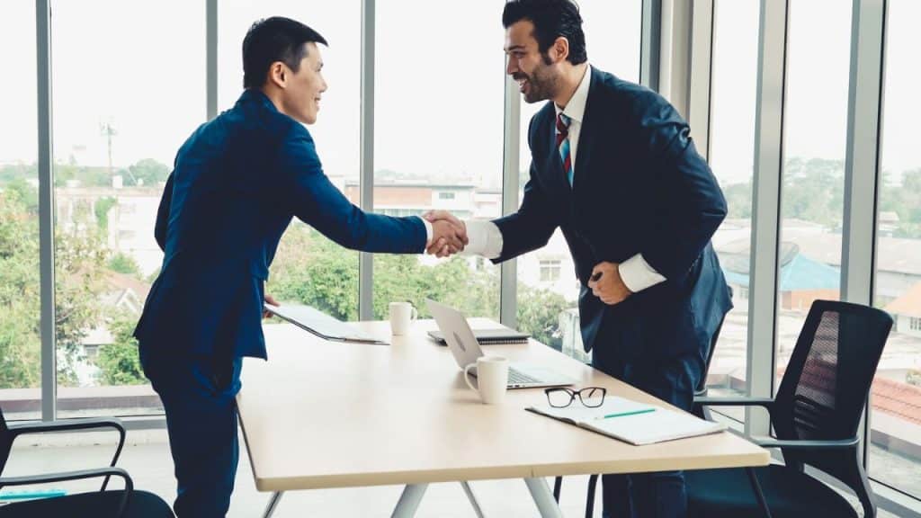 Two men in suits are shaking hands across a table in an office setting.