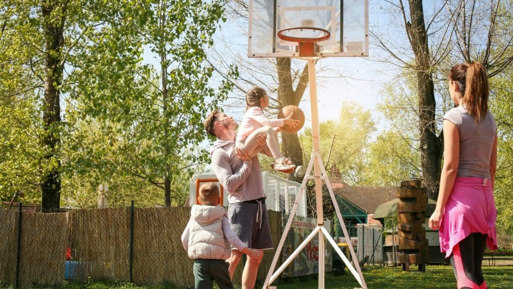 A father lifting his daughter toward an outdoor basketball hoop.