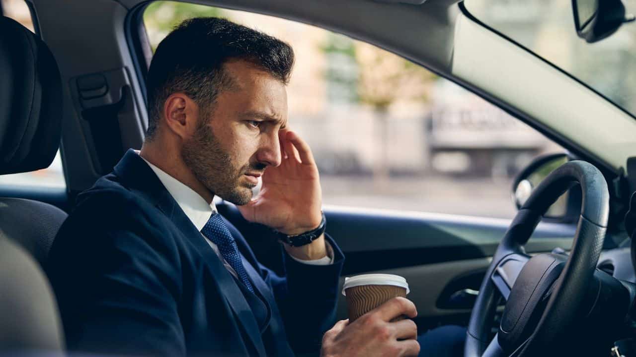 A distressed man in a suit sits in a car, holding a coffee cup.