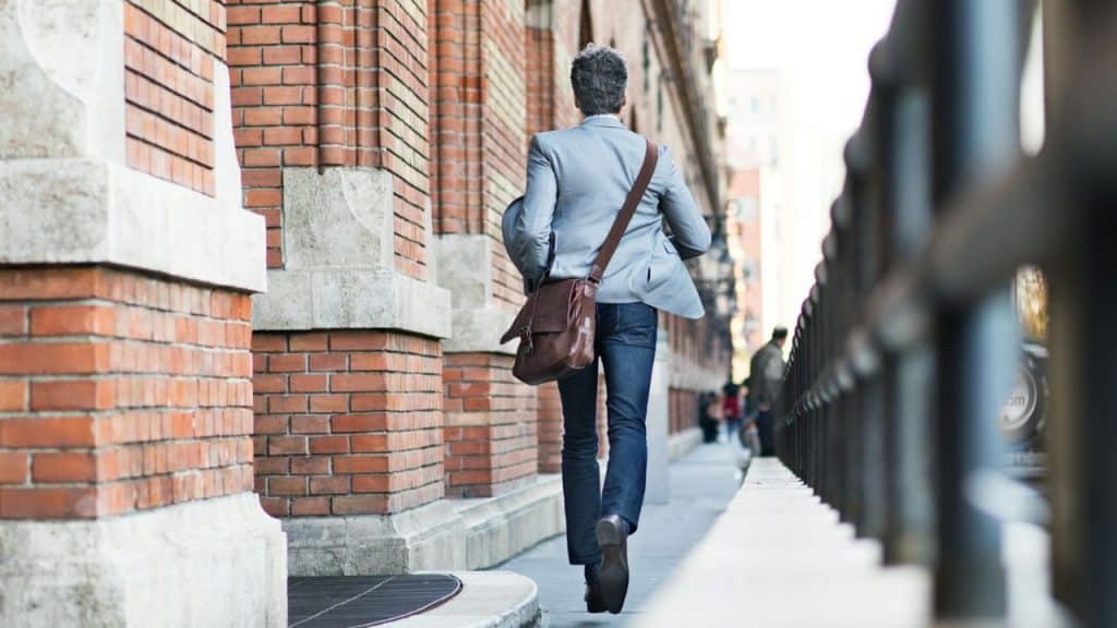 A man with a satchel walks away from the camera down a city sidewalk.