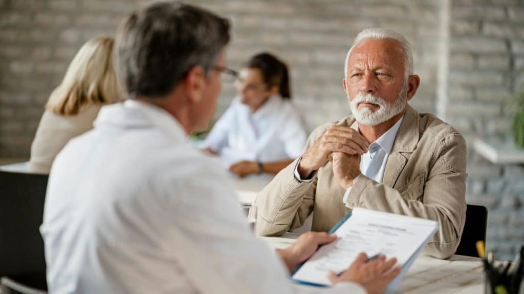 A bearded man in a tan jacket sits across from a doctor in a white coat, discussing paperwork.
