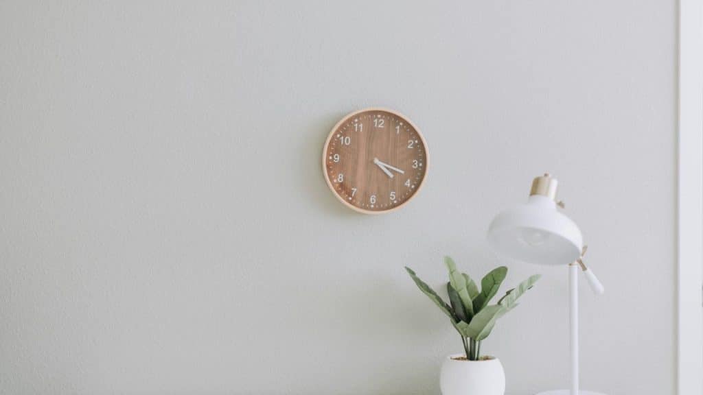 A wooden wall clock hanging above a white desk lamp and a small potted plant.