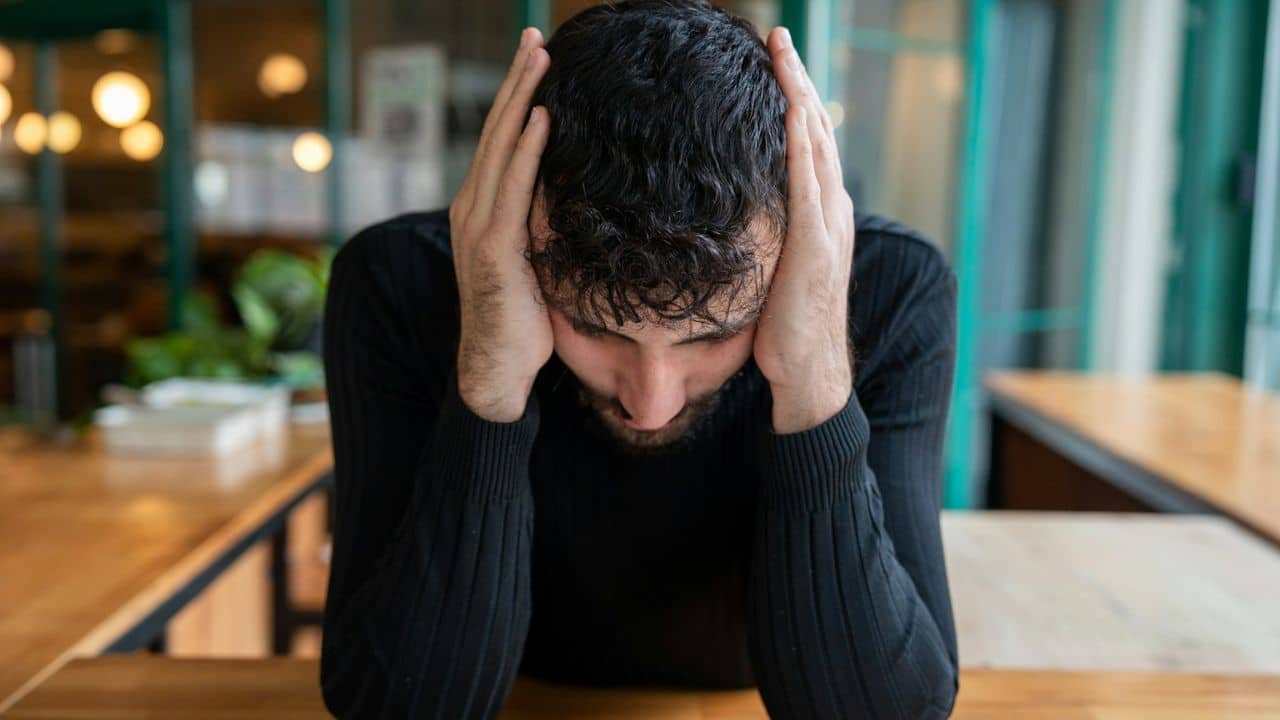 A man with dark, curly hair holds his head in his hands, looking down at a wooden table.