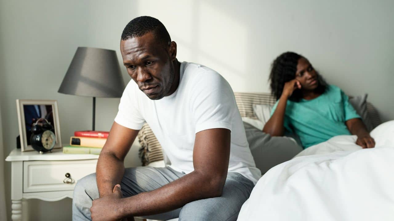 A man in a white t-shirt sits dejectedly on the edge of a bed, while a woman in a green shirt looks away in the background.