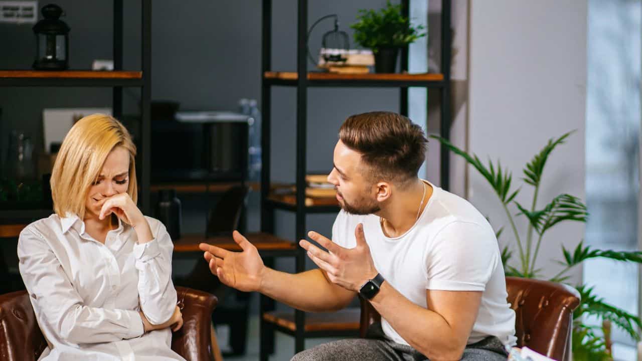 A woman with a pained expression sits beside a man who is arguing, gesturing with his hands.