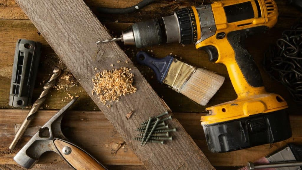 A yellow cordless drill, paintbrush, screws, and drill bits arranged on a wooden plank.