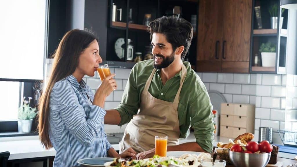 A smiling man in an apron watches a woman drink juice in a kitchen.