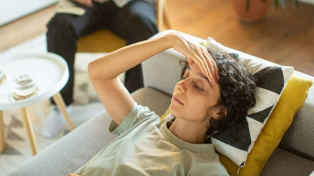 A woman with curly hair lies on a couch, hand on her forehead, eyes closed.