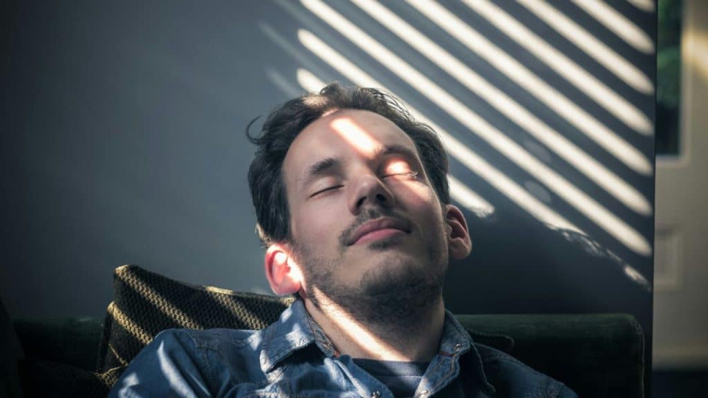 A man with closed eyes rests his head back, with light stripes on his face.