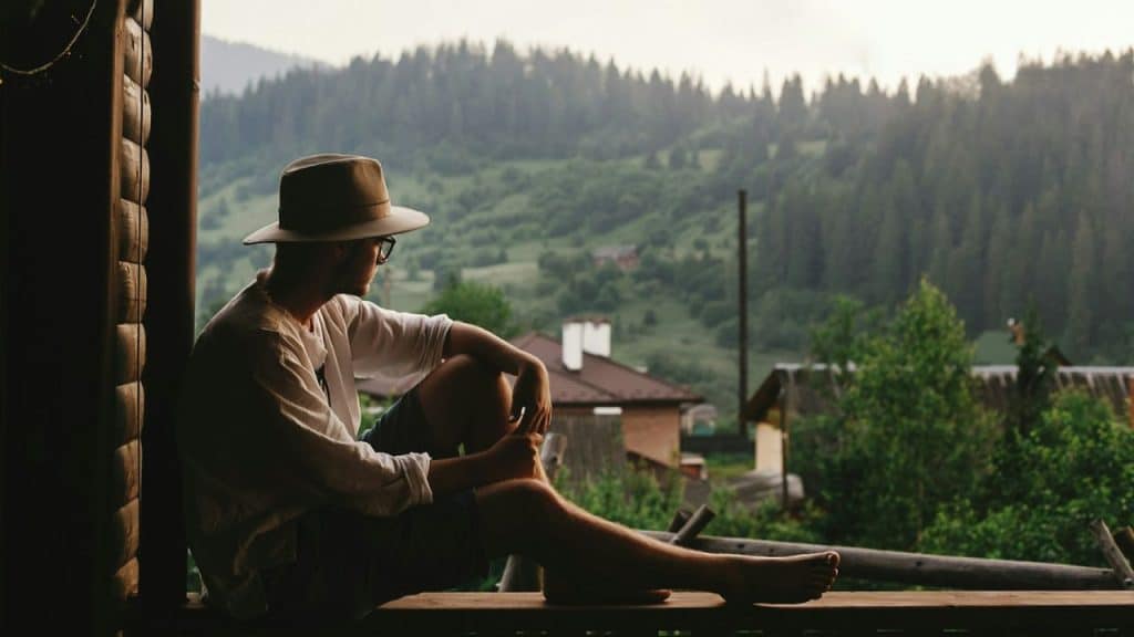A man in a hat and glasses sits on a porch, looking out at a mountain landscape.