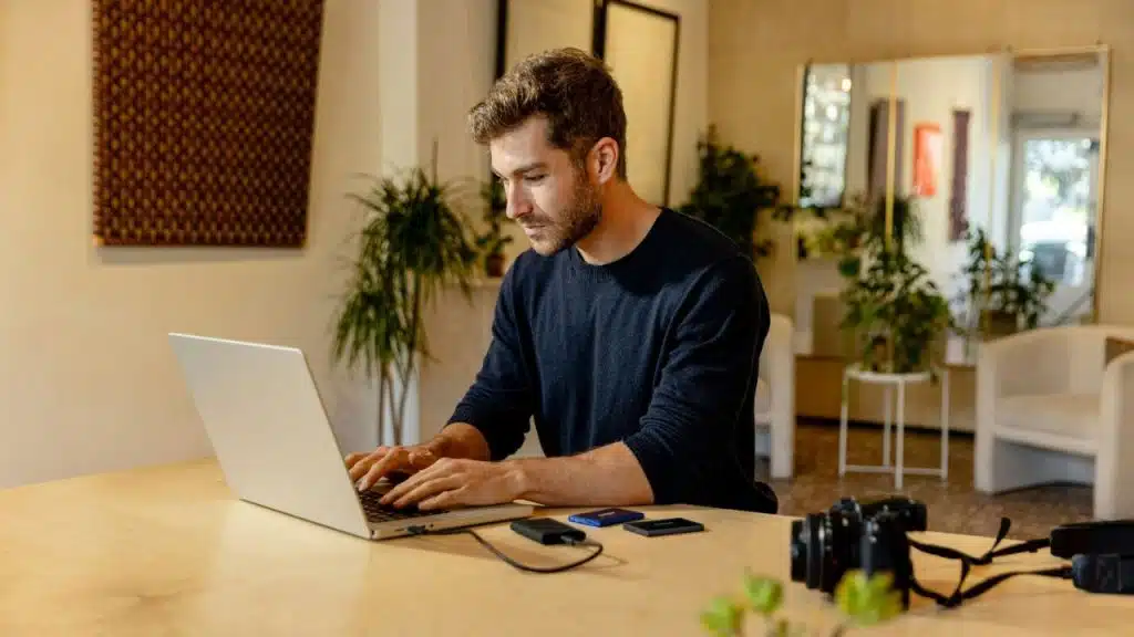 A man with a beard sits at a wooden table, typing on a laptop. There's a camera and other items on the table.