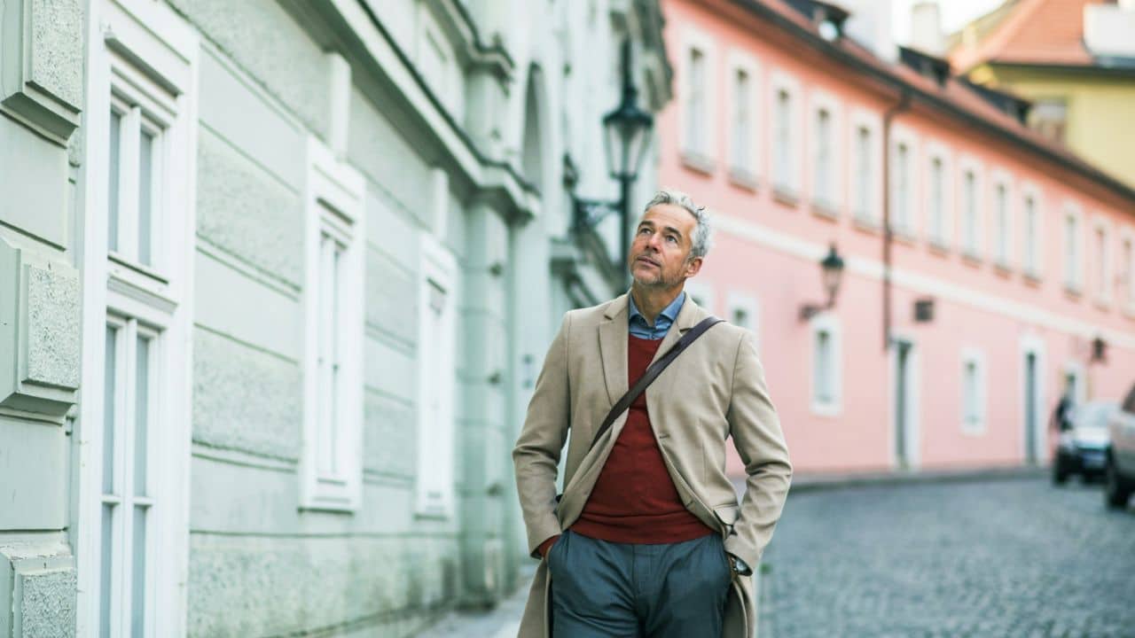 A grey-haired man in a beige coat walks on a cobblestone street, looking up.