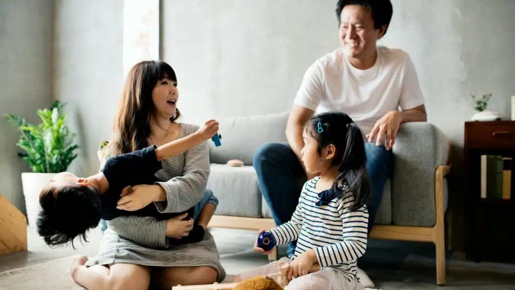 A happy Asian family plays together in a living room; a woman holds a child, and another child sits on the floor with toys, while a man sits on a sofa, looking at his child with a smile on his face.