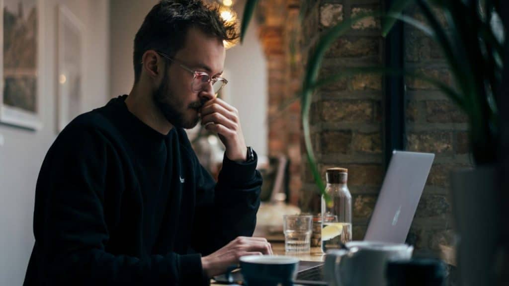 A man with glasses and a beard sits at a table, looking at a laptop.
