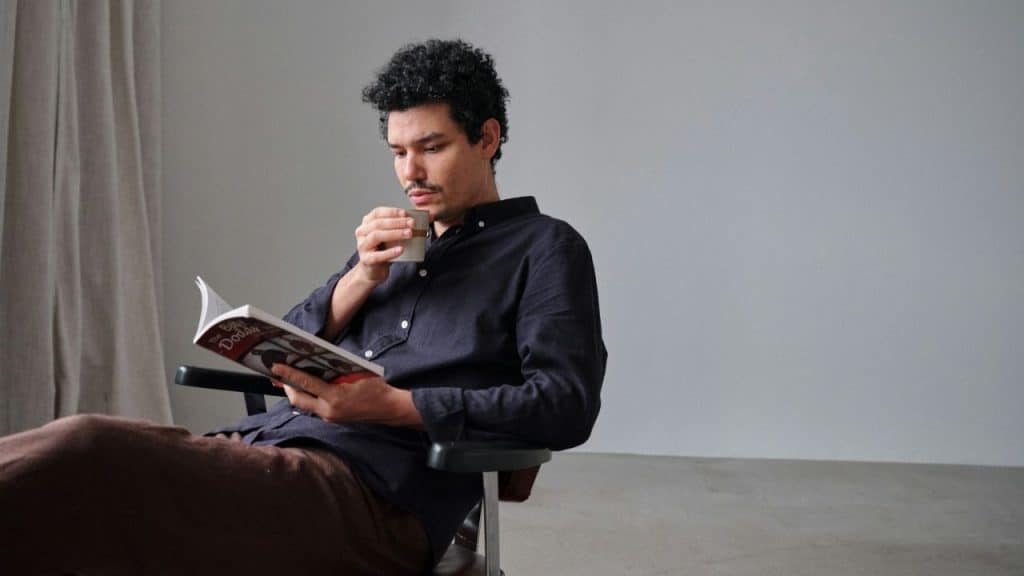 A man with curly hair and a mustache reads a book while holding a cup.