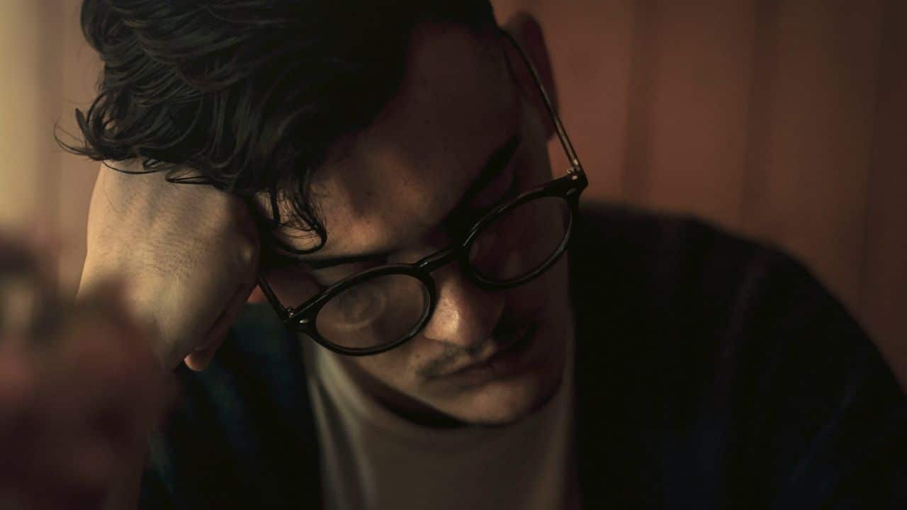 A man wearing round glasses rests his head on his hand, looking down.