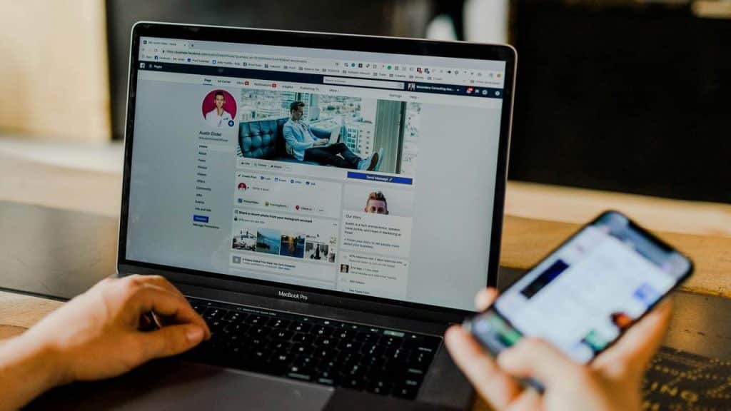 A person using a laptop displaying a Facebook business page while holding a smartphone.