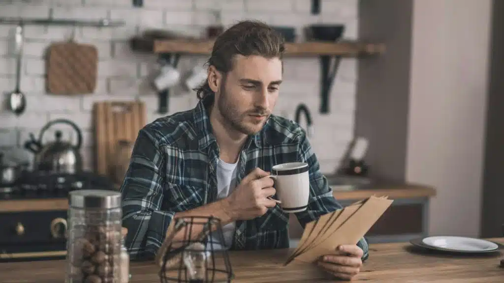 A man in a plaid shirt sits at a kitchen counter, holding a mug and looking at papers.