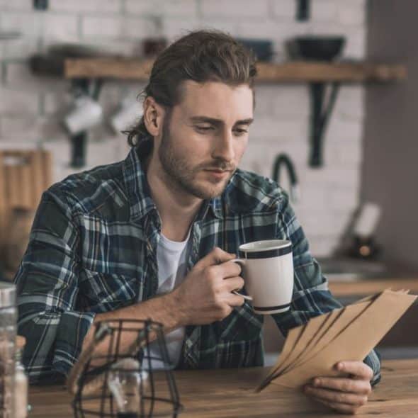 A man in a plaid shirt sits at a kitchen counter, holding a mug and looking at papers.