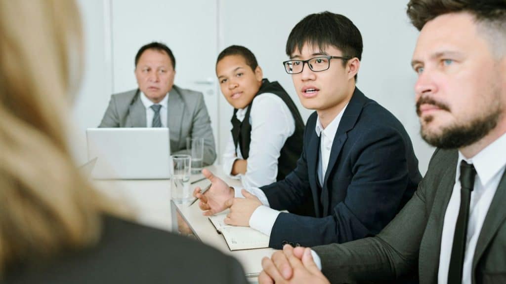 A young man in a suit and glasses speaks at a table with other people.
