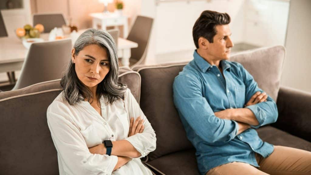 A disgruntled couple sits with arms crossed on separate couches, looking away from each other.