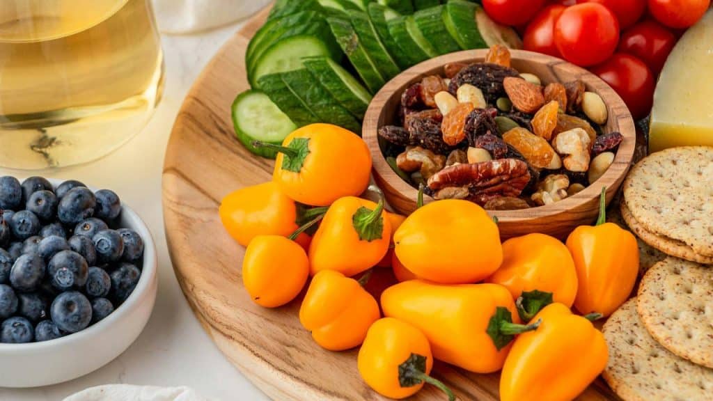 A snack board with mini yellow peppers, mixed nuts, crackers, cheese, blueberries, and cucumber slices.