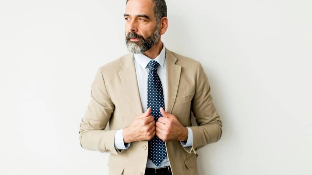 A bearded man in a beige suit adjusting his tie.