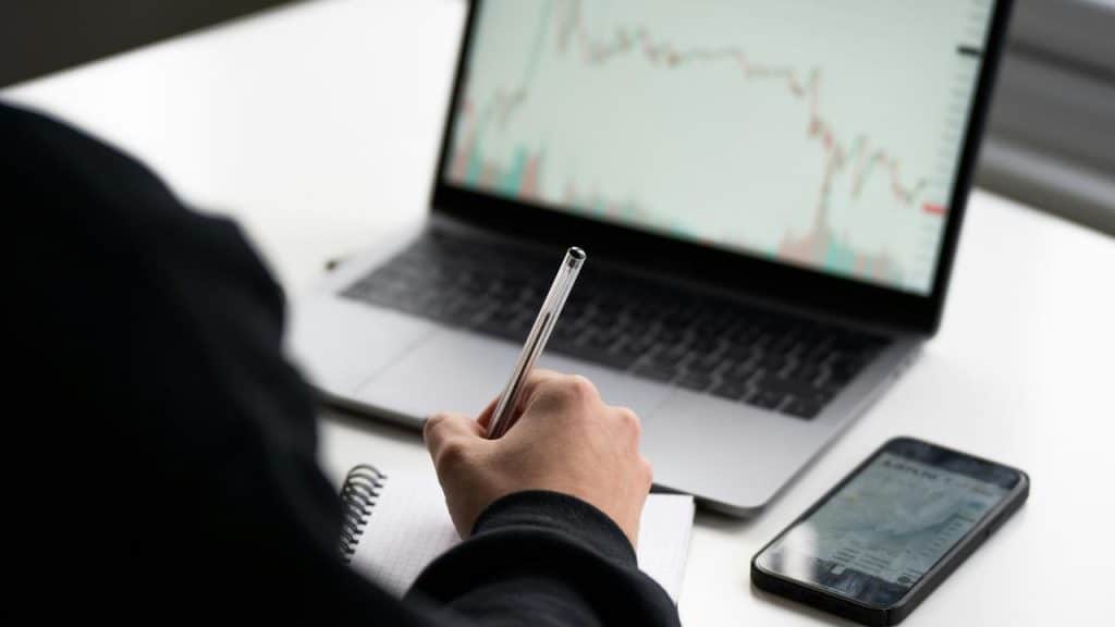 A person viewing stock charts on a smartphone in front of a laptop.