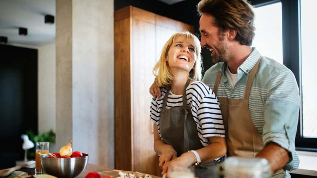 A happy couple laughs and embraces while cooking in a sunlit kitchen.