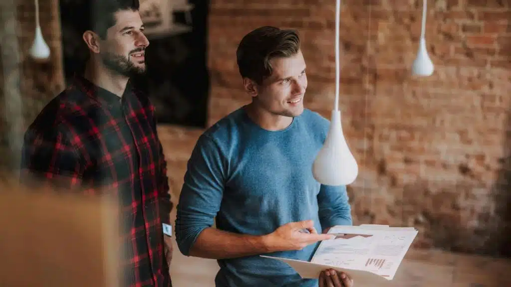 A man pointing at charts in a folder while another looks on.