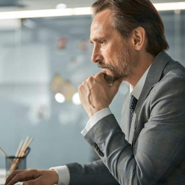 A serious man in a suit looks at a laptop, hand on chin.