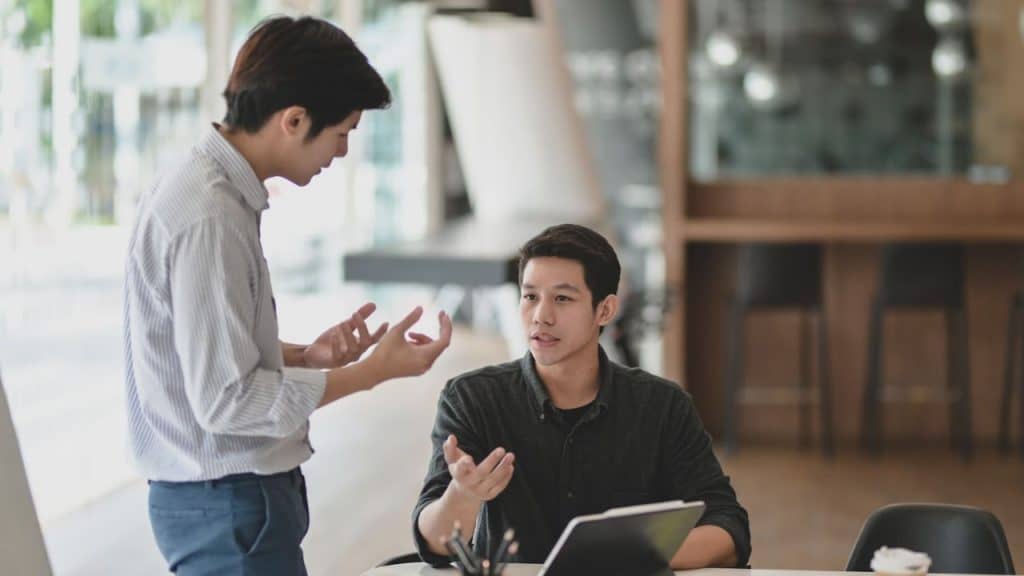 Two men are talking in an office, one standing and gesturing, the other seated and engaged.