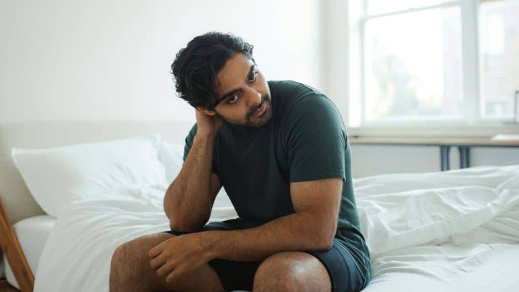 A disheveled man sits on a bed, looking off to the side, with hand on neck.